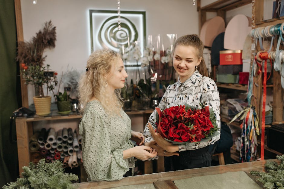 A young woman with light brown hair tied back, smiling, receives a vibrant bouquet of red roses, red hypericum berries, and green foliage from a florist with curly blonde hair in a floral shop. The bo