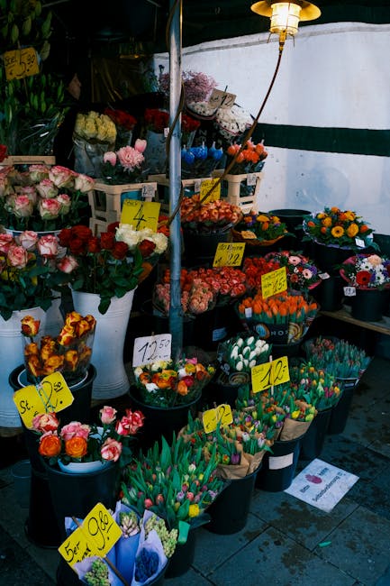 A vibrant outdoor flower market display featuring an array of fresh flowers including pink roses, orange tulips, yellow daisies, purple chrysanthemums, and multicolored tulips arranged in black and wh