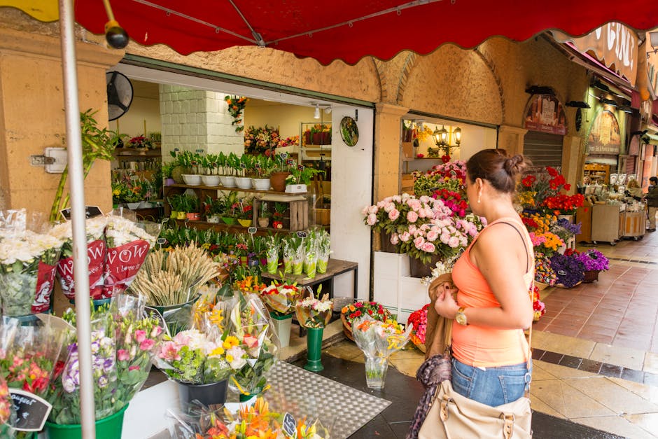 A woman with her hair tied back, wearing an orange sleeveless top and blue jeans, stands in front of a flower stall at a market. She is holding a small bouquet of flowers wrapped in brown paper in her