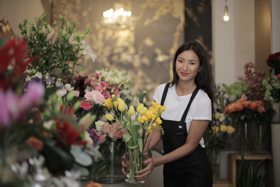 A young woman with long dark hair, wearing a white t-shirt and black apron, is smiling while holding a clear glass vase filled with a floral arrangement of yellow tulips and small white flowers. She i