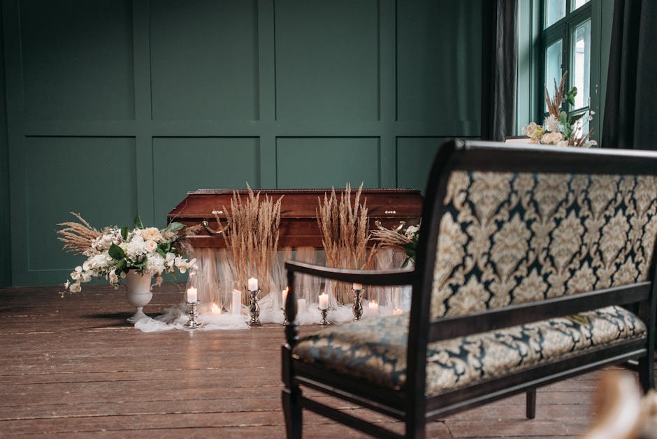 A floral arrangement with soft white roses and delicate greenery is displayed in a white ceramic vase on a dark wooden table, accented by tall, dried pampas grass and smaller white candles in silver h