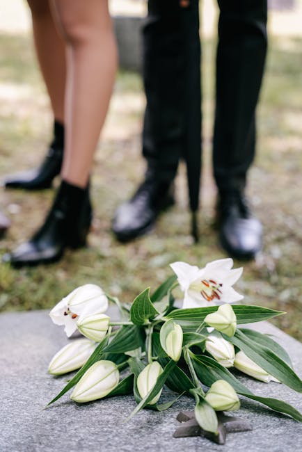 A close-up view of a bouquet of white lilies with partially opened blooms and green leaves, resting on a stone surface outdoors. In the background, two people are standing, with only their legs visibl