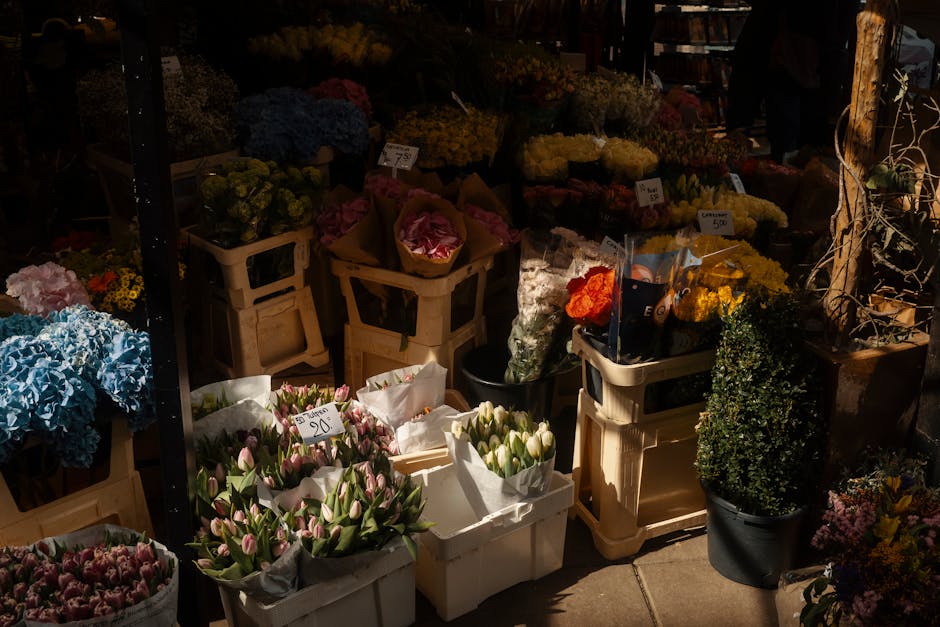 A vibrant display of fresh flowers arranged in baskets and crates at a flower market stall. There are clusters of pink and white tulips wrapped in white paper, with some yellow and blue hydrangeas nea