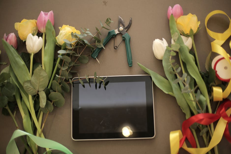 A flat-lay arrangement of fresh tulips in shades of pink, yellow, and white, interspersed with green eucalyptus leaves, surrounds a tablet device placed in the center. The flowers are arranged with so