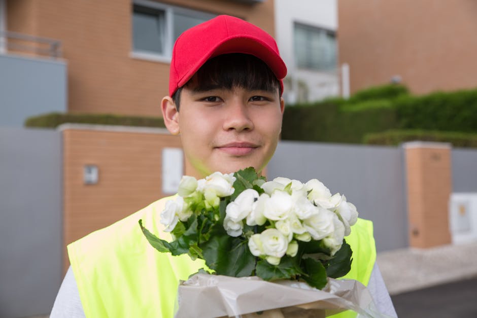 A young male courier wearing a red cap and high-visibility yellow vest holding a bouquet of fresh white flowers wrapped in paper. The flowers include white roses and other white blossoms with green le