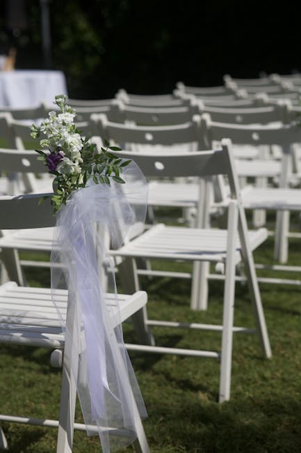 A floral arrangement attached to a white wooden chair at an outdoor wedding setting. The arrangement features a small bouquet of white and purple flowers, including lisianthus and other delicate bloom