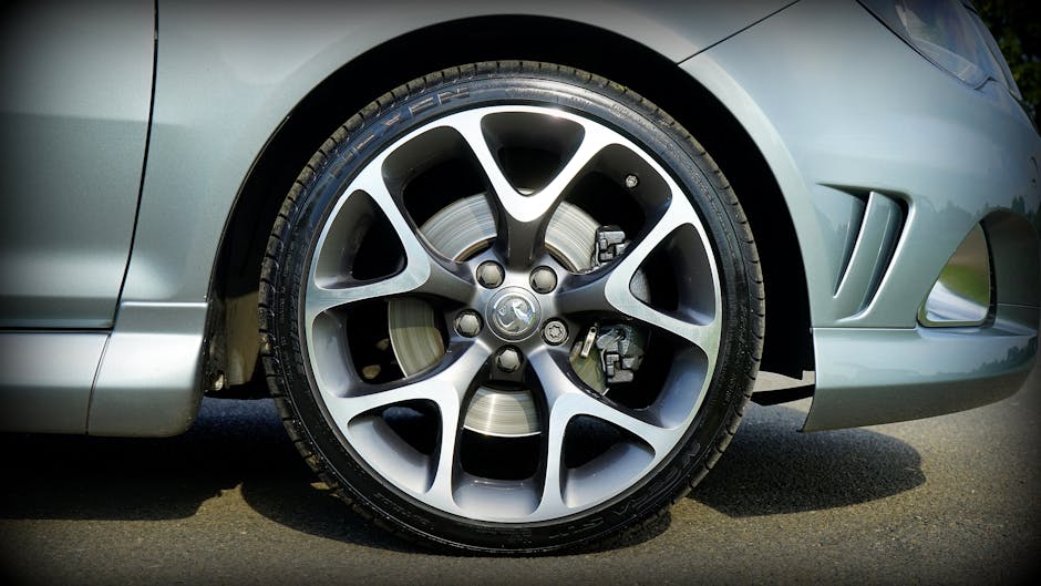 Close-up of a silver car's front wheel, featuring a modern alloy rim with black and silver spokes, a visible brake disc and caliper behind the rim, set on an asphalt road with sunlight reflecting off
