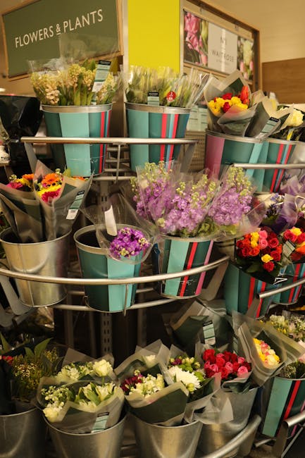 A display of fresh flowers arranged in various small metal and striped paper buckets on a multi-tiered metal stand inside a florist shop. The bouquets include a mix of colorful roses, purple statice,