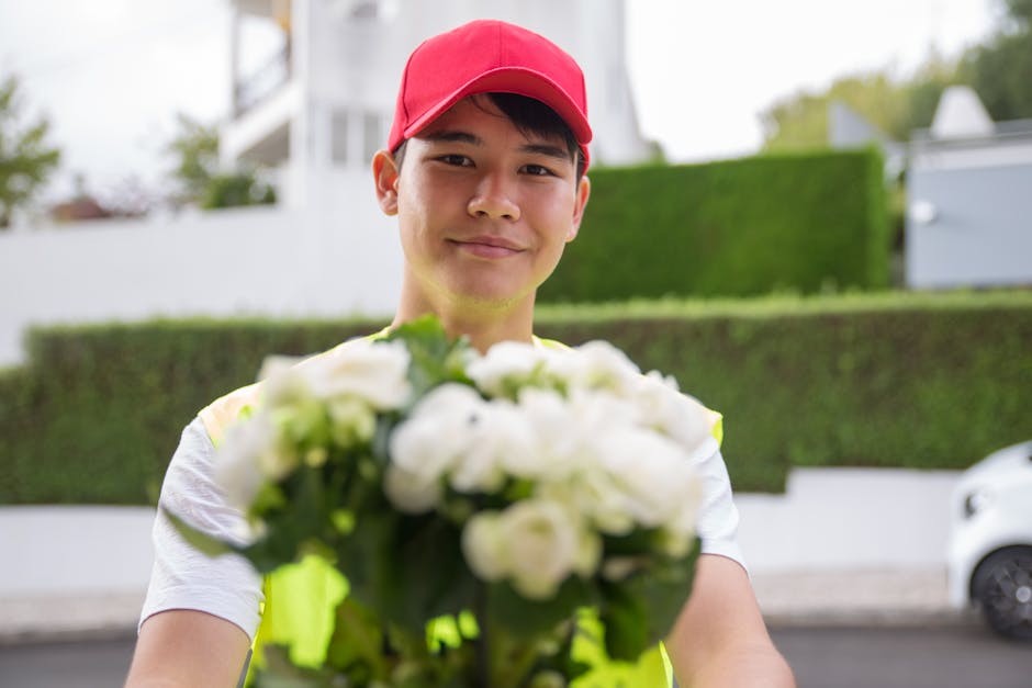 A delivery person wearing a red cap and high-visibility vest is holding a bouquet of fresh white flowers, including roses and peonies, wrapped in clear cellophane. The arrangement is lush and well-str